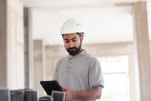 Bearded contractor in hard hat reviewing plans on a tablet at a job site