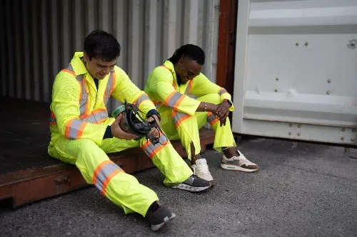 Two exhausted workers in hi-vis suits resting against a shipping container