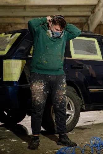 Auto body worker in respirator mask with paint-covered clothing in garage
