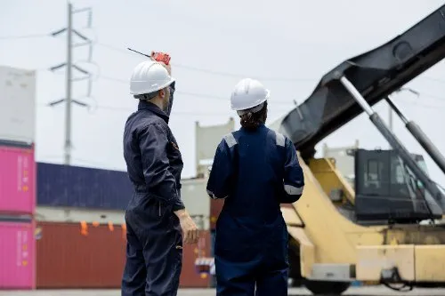 Two workers in hard hats overseeing shipping containers at port
